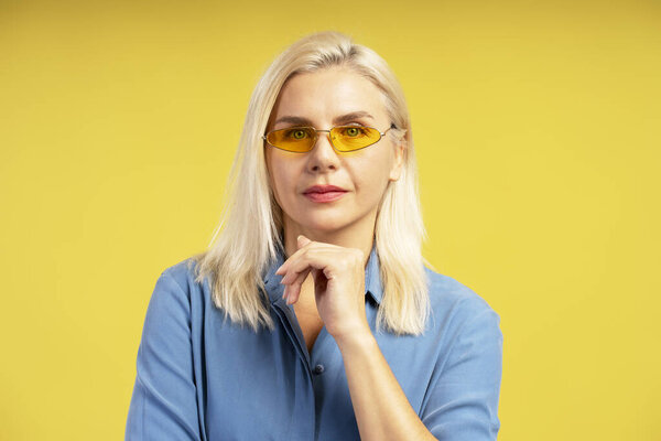 Studio portrait of a blonde businesswoman wearing yellow sunglasses and a blue shirt, posing with her hand on her chin on a vibrant yellow background