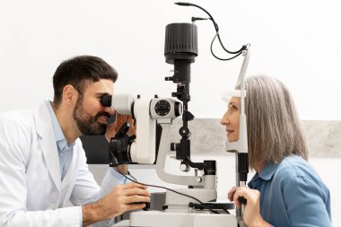Modern bir klinikte biyomikroskop kullanarak vizyon ve göz sağlığını değerlendiren kıdemli bir kadın hastanın gözlerini inceleyen oftalmolog. Sağlık hizmeti kavramı