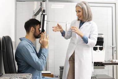 Kıdemli bayan göz doktoru bir erkek hasta üzerinde fosforopter kullanarak göz muayenesi yapıyor, lensleri ayarlıyor ve modern bir göz kliniğinde ona yol gösteriyor. Tıbbi konsept