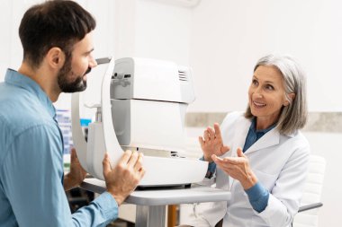 Göz muayenesi sırasında phoropter kullanan bir oftalmolog, modern bir oftalmoloji kliniğindeki bir erkek hastaya prosedürü açıklıyor ve muayene boyunca rahatlık ve anlayış sağlıyor. Sağlık hizmeti kavramı