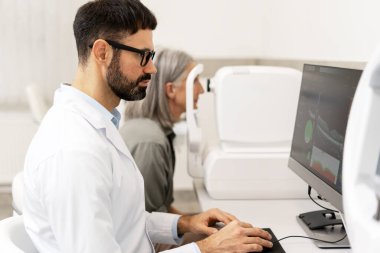 Optimetrist, modern bir göz hekimliği kliniğinde bir bilgisayarın görme testinin sonuçlarını analiz ederken, hasta arka planda kapsamlı bir göz muayenesinden geçiyor.