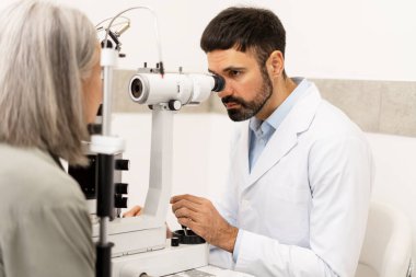 Doctor using medical equipment and performing eye exam on mature patient, checking vision and diagnosing potential problems, providing professional healthcare in ophthalmology clinic clipart