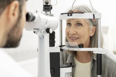 Kesik lamba kullanan bir optometrist, rutin bir göz kontrolü sırasında gülümseyen yaşlı bir kadının gözlerini incelemek, optimum görüş sağlığını sağlamak ve genel olarak refahı arttırmak