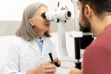 Üst düzey kadın göz doktoru, modern bir klinikte kapsamlı bir göz kontrolü sırasında doğru reçeteyi belirlemek için lensleri ayarlayan bir fosforopter kullanarak erkek bir hastanın görme yeteneğini inceliyor.