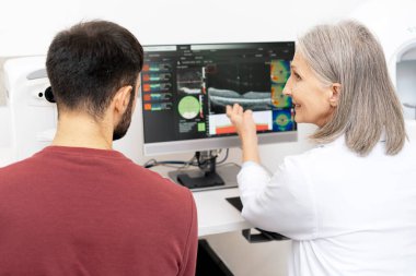 Kıdemli kadın göz doktoru, bir göz hekimliği kliniğinde tıbbi danışma sırasında bilgisayar ekranındaki sonuçları ve bunları bir erkek hastaya açıklıyor.