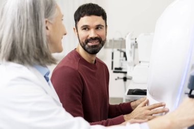 Optometrist explaining eyesight test procedure to a smiling male patient sitting next to a modern ophthalmology equipment in her bright clinic clipart