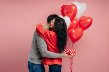Romantik çift şefkatle kucaklaşıyorlar, ellerinde kalp şeklinde balonlar ve bir hediye, Sevgililer Günü 'nde aşklarını pembe bir fon ile kutluyorlar, Sevgililer Günü konseptiyle.