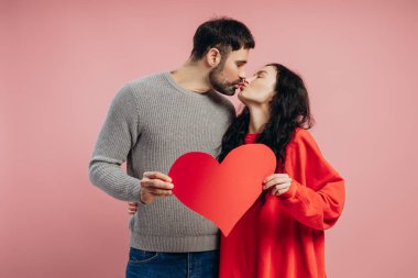 Genç çift büyük kırmızı bir kalp tutarken tutkuyla öpüşüyor, sevgililer gününü canlı pembe bir arka plana karşı kutluyor, aşk ve romantizm saçıyor. Sevgililer Günü konsepti