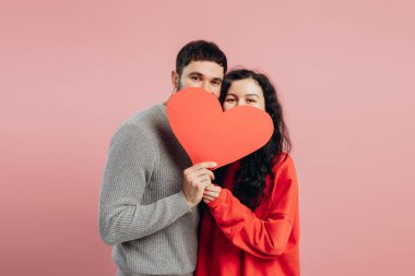 Mutlu çift büyük kırmızı bir kalp taşıyor, yüzlerini saklıyorlar ve gülümsüyorlar, sevgililer gününü pembe arka planda kutluyorlar, aşkı ve sevgiyi ifade ediyorlar, romantik anları paylaşıyorlar. Sevgililer Günü konsepti