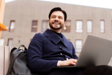 Şehirde bankta oturmuş, dizüstü bilgisayarla çalışan gülümseyen, serbest çalışan genç bir erkek. Tarafını arıyor. İş, ağ, teknoloji kavramı