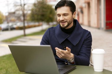 Paltolu, gülümseyen genç bir adamın portresi. Kafede oturmuş, internette sohbet ediyor, dizüstü bilgisayar kullanıyor. İş, ağ, teknoloji kavramı