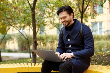 Sırt çantalı gülümseyen sakallı genç adam dizüstü bilgisayarda çalışıyor, şehirdeki parkta oturuyor. İş, ağ, teknoloji kavramı