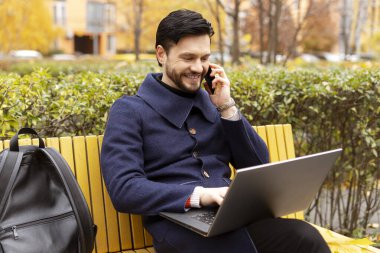 Akıllı telefonda konuşan mutlu sakallı adam dizüstü bilgisayarla yazarken, şehir parkında bankta otururken. İş, ağ, teknoloji kavramı