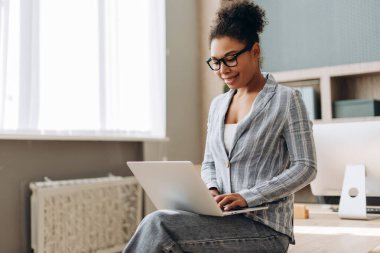 Genç yönetici bir masada oturuyor, dizüstü bilgisayarla yazı yazıyor ve modern bir ofiste bilgisayarla gülümsüyor ve arka planda bir pencerenin yanında raflar var.