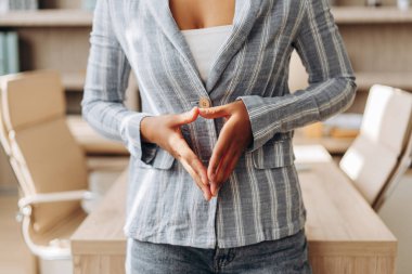 İş kadını şık bir spor ceket giyiyor, modern bir ofiste güvenle duruyor, parmaklarıyla üçgen el hareketi yapıyor, odak, bağlantı ve yaratıcılığı sembolize ediyor.