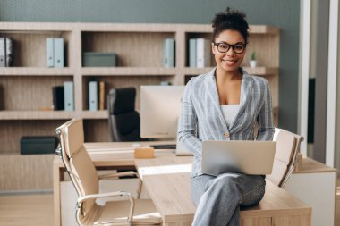 İş kadını, modern bir ofiste dizüstü bilgisayarla masasında otururken gülümsüyor. Çalışma alanında kendine güvenini ve profesyonelliğini gösteriyor.