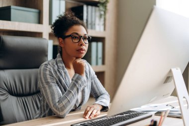 Orta yaşlı Afrikalı Amerikalı kadın, masa başında oturan ve çenesine dokunan iş kadını modern ofiste arka planda kitaplığı olan bilgisayar monitörüne dikkatlice bakarken. İş konsepti