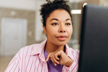 Elleri çenesinde genç bir ofis çalışanı, düzgün, modern ofis ortamında bir masaüstü bilgisayarıyla uğraşırken dikkatle konsantre oluyor.