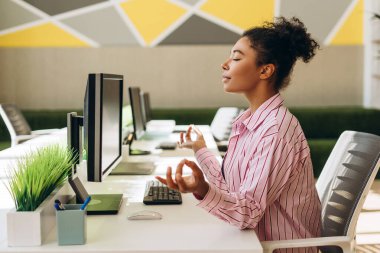 Genç iş kadını, modern ofisindeki bilgisayar masasında farkındalık meditasyonu yapıyor, iş yerindeki zihinsel sağlık ve stres azalmasını teşvik ediyor.