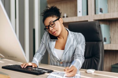 Odaklanmış kadın yönetici, parlak, modern bir ofiste birden fazla iş yapıyor, aynı anda bilgisayar üzerinde çalışıyor, telefonla konuşuyor, notlar alıyor, iş yükünü verimli bir şekilde yönetiyor. İş konsepti