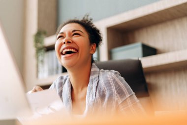 Genç iş kadını bir belgede heyecan verici haberler okurken gülüyor, ofis masasında başarıyı kutluyor, mesleki çevresinden neşe ve memnuniyet yayıyor.