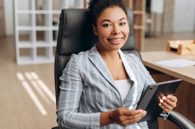 İş kadını rahat bir şekilde ofis koltuğunda oturuyor, elinde dijital bir tablet tutuyor ve gülümsüyor, muhtemelen raporları gözden geçiriyor veya toplantıya hazırlanıyor.