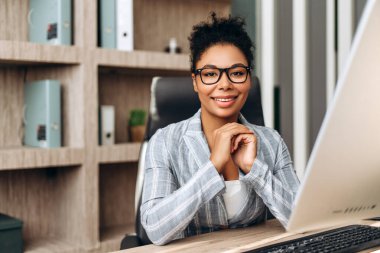 Kendine güvenen genç yönetici elleri sımsıkı bağlanmış ve gülümsüyor, modern ofiste bilgisayarda çalışıyor ve arka planda dizinlerle dolu raflarla dolu.