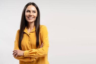 Sarı tişörtlü genç bir iş kadınının stüdyo portresi, kollarını kavuşturup gülümseyen ve diğer tarafa bakan, özgüvenini ve profesyonelliğini beyaz bir arka plana yansıtan