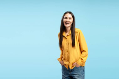 Uzun kahverengi saçlı, sarı tişörtlü ve kot pantolonlu neşeli bir kadının stüdyo fotoğrafı. Elleri rahat bir şekilde cebine sokulmuş, canlı mavi bir arka planda.