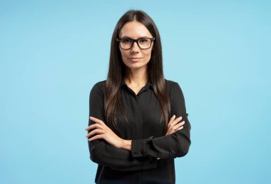 Gözlüklü ve siyah gömlekli bir iş kadını portresi, kolları çapraz mavi bir arkaplanda duruyor, özgüven ve profesyonellik yayıyor. İş konsepti