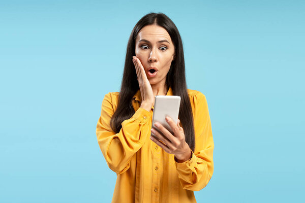 Shocked Latin businesswoman is using mobile phone, reading shocking news, touching face with hand, standing on blue studio background, expressing astonishment and surprise
