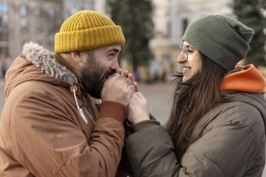 Soğuk bir kış gününde, şehir ortamında ellerini ısıtan, romantik bir anın tadını çıkaran ve sıcaklığı ve sevgiyi paylaşan gülümseyen çift.