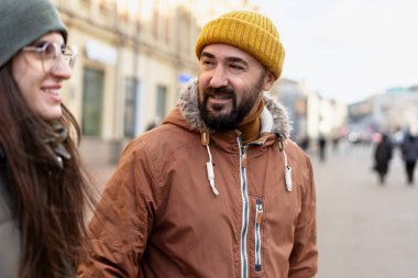 Sakallı bir adam, bereli ve kışlık ceketli. Kışın sokakta yürürken gözlüklü ve bereli bir kadınla gülümsüyor ve konuşuyor.