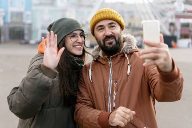 Kış kıyafetleri giymiş neşeli turistler, canlı bir lunaparkta birlikte vakit geçirirken video görüşmeleri yaparken mutlu bir şekilde kameraya el sallıyorlar.