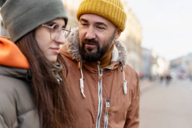 Sarı bir bere takan sakallı baba, kışın serin bir şehir sokağında birlikte dikilirken, kışın giyinmiş, üzgün genç kızını teselli ediyor.
