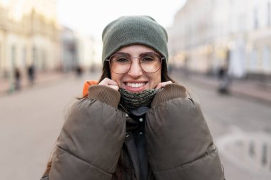 Gözlüklü, kalın bir şapka ve yastıklı ceket takan neşeli genç bir kadın, soğuk bir kış günü şehir caddesinde yürürken gülümseyerek eşarbını düzeltiyor.