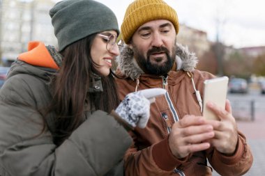 Kışın giyinmiş iki turist, bir şehir caddesinde harita uygulamasını tartışırken akıllı bir telefonla serin şehir manzarasında geziniyorlar.