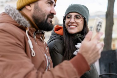 Kışlık giysiler içinde mutlu bir çift, dışarıda akıllı bir telefonla neşeli bir selfie çekiyorlar, soğuk bir kış gününün sıcaklığını ve eğlencesini kucaklıyorlar.