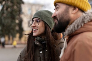 Sıcak örülü şapkalar ve kalın kışlık ceketler giyen genç bir çift, gülümseyerek ve şehirde keyifli bir günün tadını çıkararak, serin mevsimlerde birlikte olmanın keyfini çıkarıyorlar.