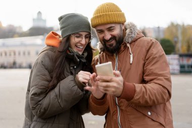 Neşeli bir adam ve kadın, rahat kışlık giysiler giyiyorlar, canlı bir şehirde, sonbahar boyunca birlikte dikiliyorlar, akıllı bir telefondan bakarken neşeyle gülüyorlar.