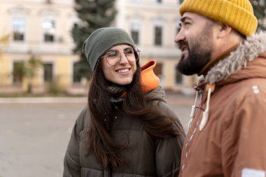 Genç bir kadın ve erkek, bulutlu bir günde sıcacık kışlık giysiler içinde birbirlerine sıcak bir gülümsemeyle dışarıda canlı bir sohbetten zevk alıyorlar. Aralarındaki bağ mutluluk saçıyor.