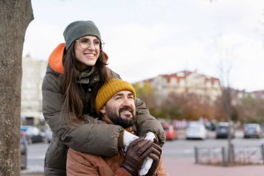 Neşeli çift, kışlık şehir sokağında romantik bir anın tadını çıkarıyor. Sıcak kıyafetler ve şapkalar giyiyorlar. Kadın, adama arkadan sarılıyor ve her ikisi de mutlu bir şekilde gülümsüyor.