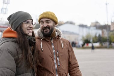 Kış kıyafetleri ve taksiler giyen mutlu çift, gülümsüyor ve arka planda bulanık binalar ve dönme dolap ile birlikte şehir meydanında bir günün tadını çıkarıyorlar.