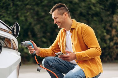 Şehirde akıllı telefon kullanarak elektrikli arabasını şarj etmek için bekleyen genç bir adamın portresi. Elektrikli araba konsepti. Yeşil seyahat