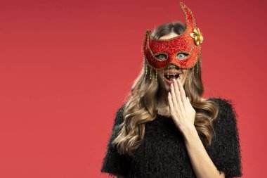 Şaşırmış bir kadın canlı kırmızı bir karnaval maskesi takıyor, elini kırmızı bir arka planda tutuyor.
