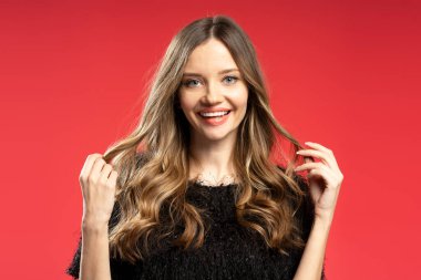 Stüdyo resmi kıvırcık saçlı, siyah kazaklı, saçlarıyla oynayan ve kırmızı arka planda gülümseyen genç bir kadın.