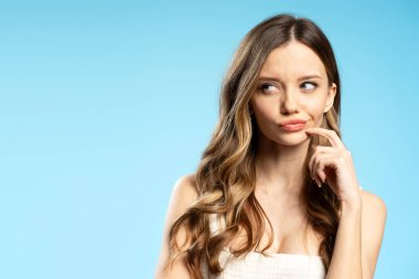 Uzun saçlı düşünceli genç bir kadının portresi mavi bir arkaplana bakarken çenesine dokunuyor.