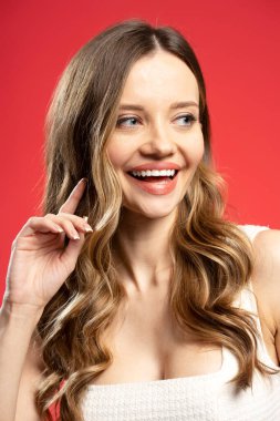 Mükemmel beyaz dişleri olan neşeli sarışın bir kadının stüdyo portresi. Saçına dokunuyor ve kırmızı arka planda gülümserken yüzünü çeviriyor. Moda konsepti