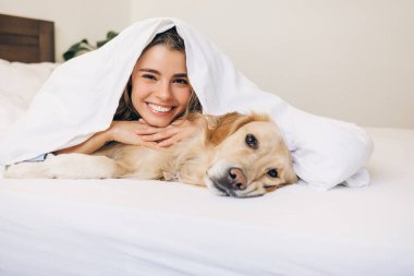 Genç kadın gülümseyen ve Golden Retriever köpeğini kucaklayan rahat beyaz bir battaniyenin altında birlikte uzanan, huzurlu ve rahatlatıcı bir sabahı evde kutlayan