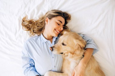 Açık mavi gömlekli gülümseyen genç bir kadın yatakta uzanmış Golden Retriever köpeğine sarılıyor. Huzurlu bir sevgi ve arkadaşlığın tadını çıkarıyor.